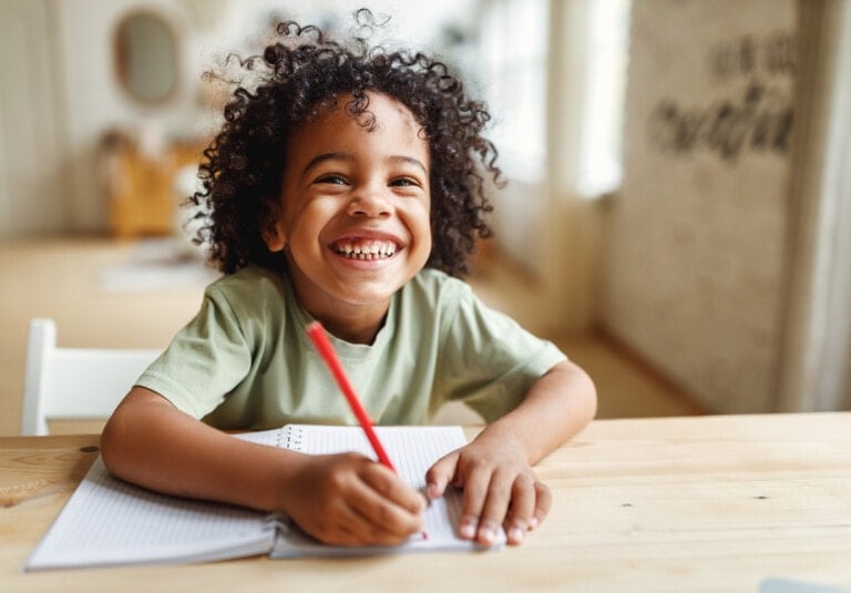 A young child with curly hair is sitting at a wooden table, smiling widely while holding a red pencil and writing in a notebook. The background is softly blurred, showing a bright, airy room with large windows and light-colored walls—an inviting space where your child learns joyfully.
