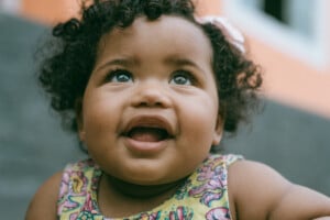 A close-up photo of a smiling baby with curly hair, possibly named one of those charming girl names that start with M. The baby is wearing a colorful sleeveless shirt with a playful pattern. The background is blurred, showcasing a building with a peach-colored wall and a white-framed window.