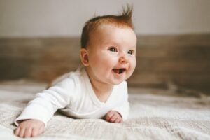 A baby with short, light brown hair and a tuft sticking up is lying on its stomach on a soft surface. The baby, potentially with one of those trendy boy names, is wearing a white long-sleeve shirt and is smiling brightly, looking slightly upward. The background is a neutral, softly blurred indoors.