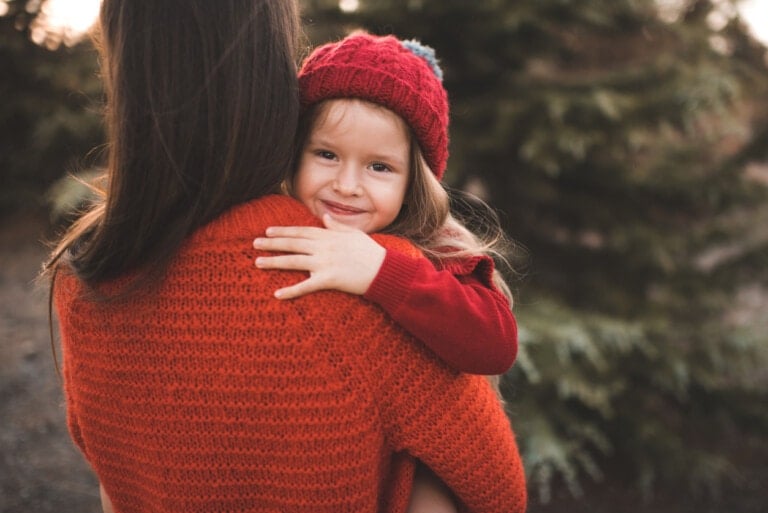 A child wearing a red beanie and a red sweater is being carried by an adult in an orange sweater. The child is smiling at the camera as they stand outdoors with blurry trees in the background, embodying hope while breaking cycles of family trauma.
