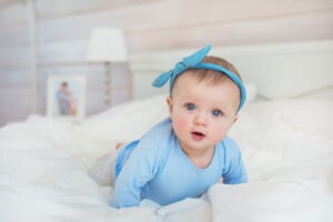 A baby wearing a blue outfit and a matching headband with a bow is lying on their belly on a white bed. The baby, possibly with one of those charming Russian girl names like Anastasia or Elena, has blue eyes and a curious expression. In the background, there is a blurred frame with a photo and a lamp.