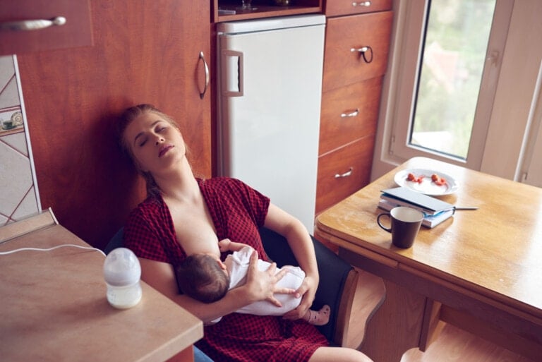 A woman is sitting in a kitchen breastfeeding an infant, appearing tired in her red checkered dress. The scene hints at her routine of triple feeding, with a baby bottle on the counter, and nearby, a table holds a plate, a notebook, a pen, and a coffee mug. A window is in the background.
