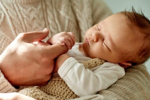 A baby sleeping peacefully in someone's arms, wearing a white outfit and a knitted blanket. The person's hands are gently holding the baby's hands. The scene exudes a sense of calm and tenderness, perfect for little ones with charming D names for boys.