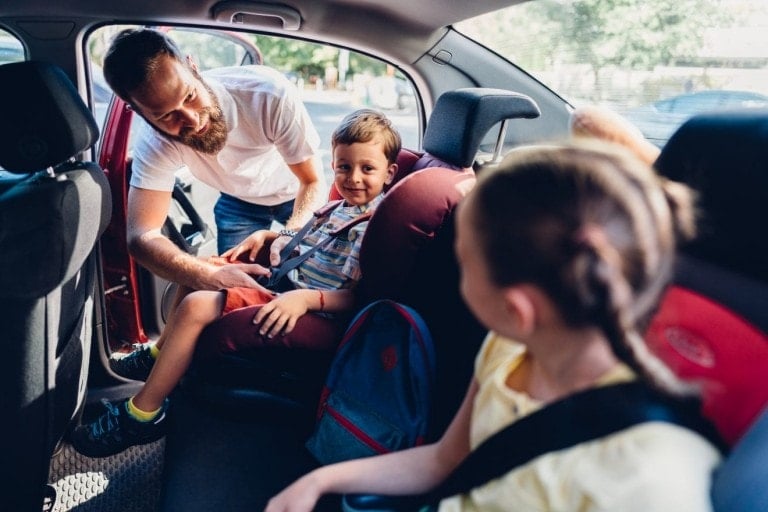 A man helps a child with a seatbelt in the backseat of a car. Another child with a ponytail and wearing a seatbelt sits in the foreground. The car's door is open, revealing trees and buildings in the background, perfect for starting their road trip activities for kids.