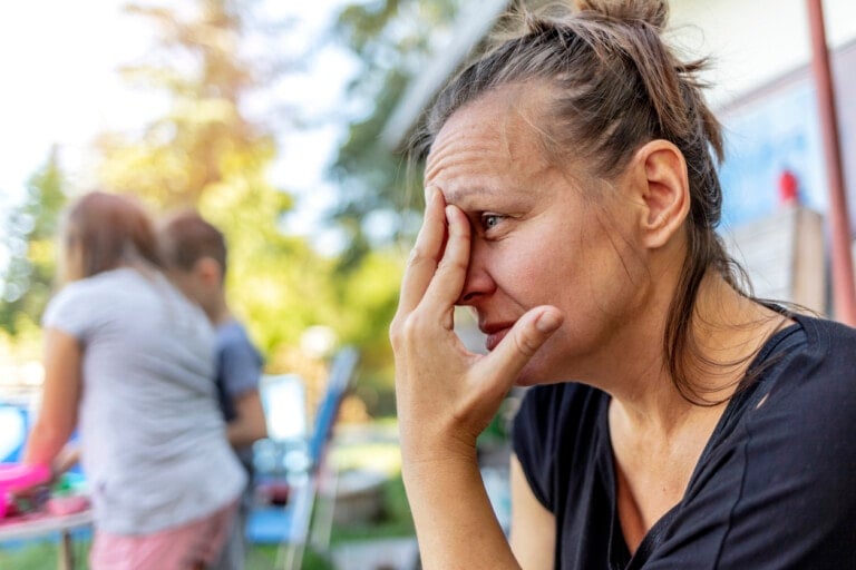 A woman is sitting outside with a hand covering part of her face, appearing stressed or tired. In the background, two children are blurred, engaged in an outdoor activity near a house. Trees and outdoor elements are visible on a sunny day, free from any unnecessary comments disrupting the scene.