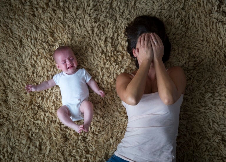 A baby in a white onesie cries while lying on a shaggy carpet, next to a woman covering her face with her hands. The woman, experiencing postpartum rage, is lying on her back in a light pink tank top and blue jeans.
