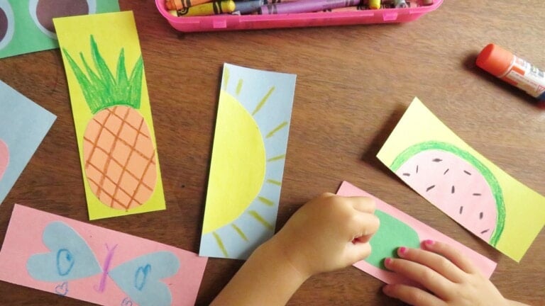 A child's hands are seen crafting on a wooden table. Various colorful paper cutouts with drawings of a pineapple, sun, watermelon, and a butterfly are spread out. A pink container with crayons, a red glue stick, and a handmade bookmark complete the vibrant scene.