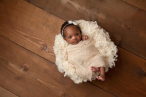 A baby wrapped in a beige blanket lies in a round, fluffy cream-colored cushion on a wooden floor. The baby girl, possibly named Talia or Tessa, is wearing a headband with a small flower and looking up at the camera.