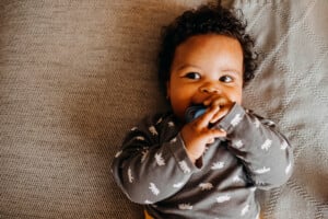 A baby with curly hair lies on a grey blanket, wearing a grey onesie with white animal patterns. The baby, possibly pondering modern boy names, holds a blue pacifier with both hands and looks slightly to the side.