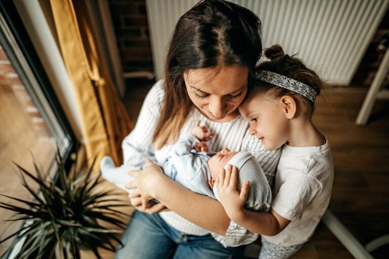 A woman holds a newborn baby in her arms while an older child, wearing a headband, gently rests their head against the baby. They are sitting in a well-lit room with a plant visible in the corner, suggesting there might be space for just one more baby.