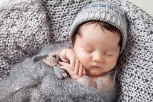 A baby named Benjamin sleeps peacefully, swaddled in a soft, gray knitted blanket. The baby is wearing a gray knit hat and has their right hand under their head. A small, crocheted stuffed toy is tucked under the baby's left arm.