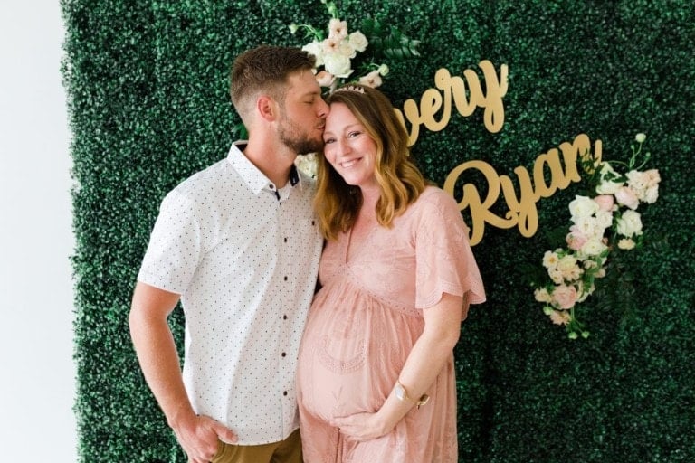 A man with short hair and a beard is kissing the forehead of a pregnant woman in a pink dress, celebrating their floral baby shower. They are standing in front of a green floral backdrop with gold text partially visible, reading "avery" and "Ryan." The woman is holding her baby bump and smiling.