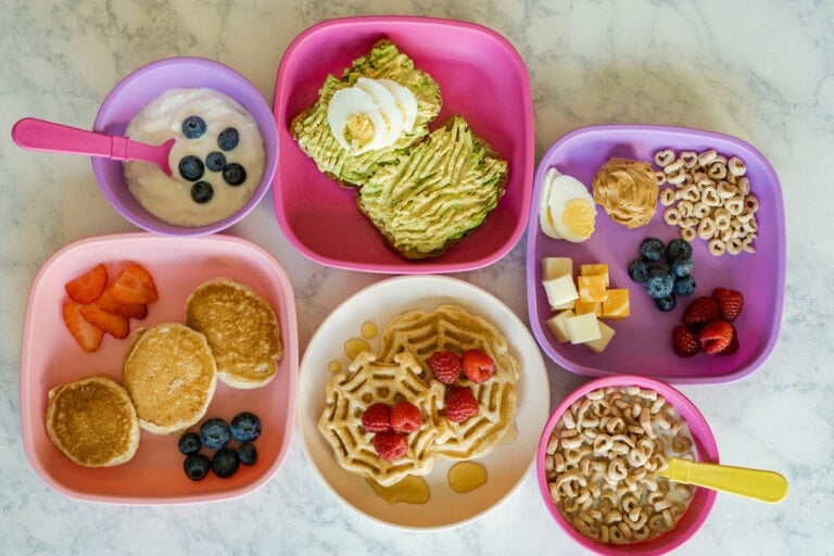 A variety of breakfast foods are served on colorful plates. Items include waffles with strawberries, avocado toast with boiled egg, yogurt with blueberries, cheese cubes with blueberries, pancakes with strawberries and blueberries, and cereal with milk—perfect healthy breakfast ideas for kids.