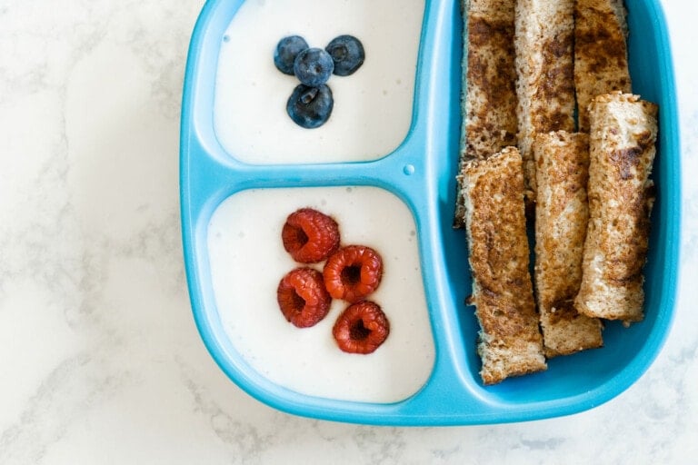 A blue divided plate holds cinnamon breadsticks on the right and two sections of yogurt on the left. The top yogurt section contains three blueberries, and the bottom section contains three raspberries. Perfect for baby-led weaning recipes, the plate rests on a white marble surface.