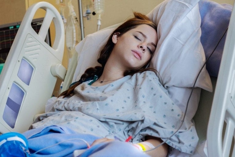 A woman with long brown hair lies in a hospital bed, wearing a hospital gown and resting with her eyes closed. She has a nasal cannula for oxygen and an IV line attached to her arm, as she undergoes labor induction. The room has medical equipment visible in the background.