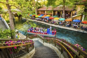 A scenic view of a river with a boat full of passengers floats by a riverside restaurant in San Antonio. Colorful umbrellas shade diners seated at outdoor tables, perfect for a babymoon outing. Flowers adorn the stairway leading down to the water, and trees and greenery create a relaxing atmosphere.