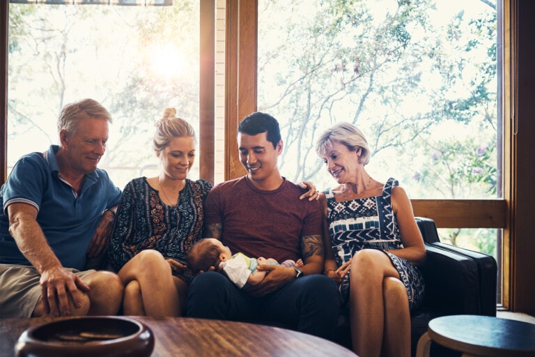 A group of five people sit on a couch, with a baby held by the person in the center. The people on either side of the person holding the baby look at the baby and smile. It’s clear they know how to set boundaries with family, creating a peaceful room with large windows and trees visible outside.