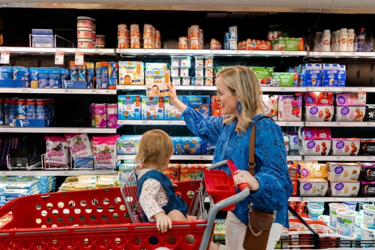 A woman in a blue blouse and white pants is shopping in a grocery store aisle with a child seated in a red shopping cart. She reaches for a box of baby formula on a shelf stocked with various baby products, including Stonyfield baby yogurt. The child gazes curiously at the products.