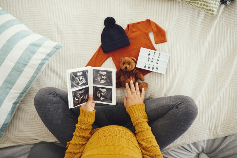 A person in their third trimester sits cross-legged on a bed, holding ultrasound images. Beside them are a small teddy bear, a baby outfit, and a sign reading "SEE YOU SOON." The bedding includes striped and patterned pillows.