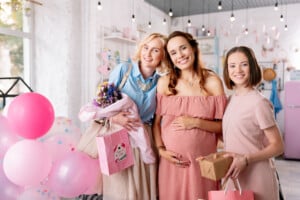 Three women are smiling indoors, with the woman in the middle visibly pregnant, cradling her belly. The other two women are holding gifts. There are pink balloons and various decorations in the background, creating a festive atmosphere with a baby shower playlist setting the mood.