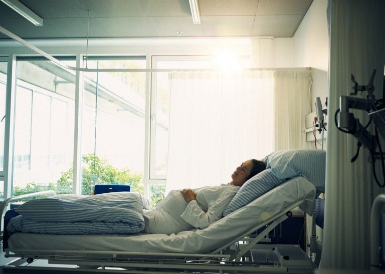 A woman lies in a hospital bed with her head slightly elevated, eyes closed, and hands resting on her abdomen. She is undergoing placenta previa treatment. The room is well-lit by natural light streaming through a large window, and medical equipment is visible on the wall next to her bed.