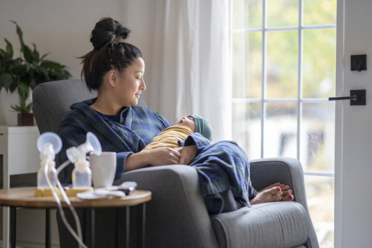 A woman sits in a chair holding a baby wrapped in a blanket, gazing out a window with natural light streaming in. On the side table next to her, there's a breast pump with an adjustable flange size, a white mug, and what appears to be a green plant in the background.