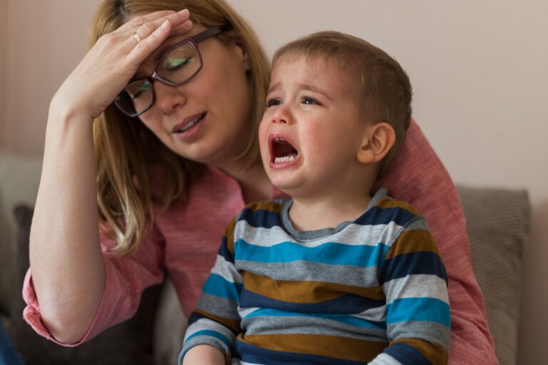 A woman holding a crying toddler in her lap appears really mad. The woman, wearing glasses and a pink shirt, has her hand on her forehead. The toddler, in a striped long-sleeve shirt, looks visibly upset. They sit on a couch with a light-colored wall in the background—an intense moment highlighting how to communicate when you're triggered.