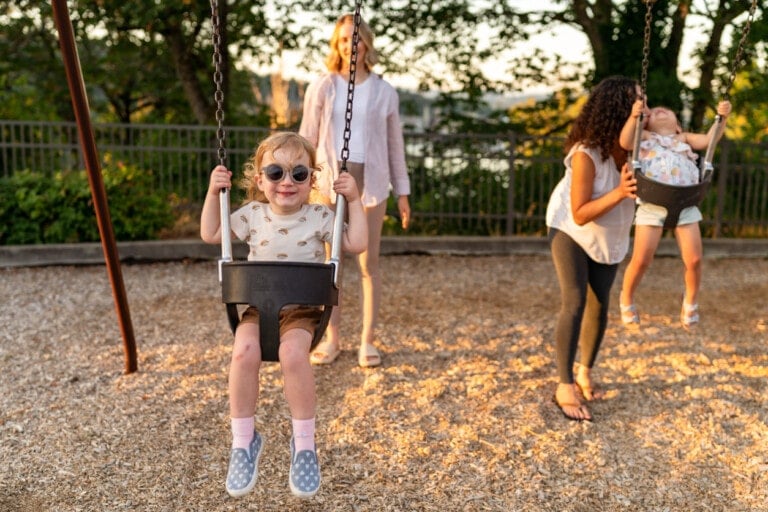 Two young children are on swings at a playground. One child in the foreground is wearing sunglasses and smiling, while the other child is being pushed by an adult woman. Another adult stands nearby, smiling. Trees and a fence are visible in the background—truly a scene of playdate must-haves.