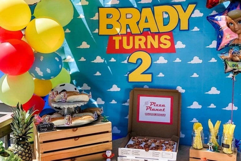 A colorful Toy Story birthday party setup with a large banner reading "Brady Turns 2" against a sky and clouds backdrop. The setup includes balloons, a wooden crate with hotdogs, an open Pizza Planet box with pizza slices, and themed decorations.