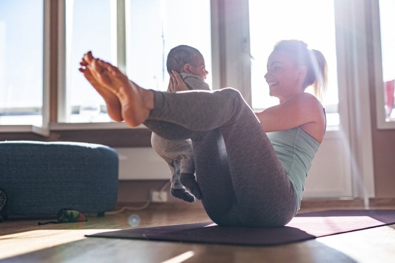 A woman in workout clothes lies on a yoga mat indoors, exercising after birth by holding a baby above her chest. She is balanced on her back with her legs bent and raised in the air, smiling at the baby. Sunlight streams through large windows in the background.