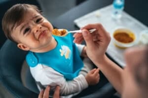 A baby in a blue bib sits in a high chair, looking displeased as an adult hand offers a spoonful of baby food. A bowl of food and a bottle are visible on the table in the background.