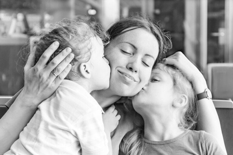 A black and white photo shows a woman mothering two young children who are kissing her on the cheeks. The woman has her eyes closed and is smiling contentedly while holding the children close with her hands. The background appears to be a cafe or restaurant.