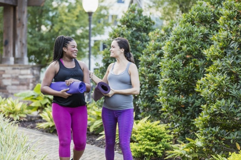 Two women walk outside in a park, holding yoga mats. One woman, wearing a dark tank top and magenta leggings, smiles at the other, who wears a gray tank top and purple leggings. They appear to be engaged in a friendly conversation about exercises you should do before giving birth amidst greenery.