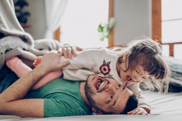 A stay-at-home dad lies on his back with a young girl playfully climbing on top of him. Both appear to be laughing and having fun. The setting is indoors, with a bright window and curtains in the background. The man is wearing a green shirt, and the girl has a floral design on her top.