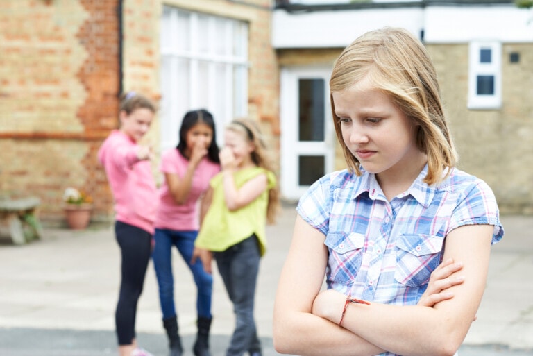 A young girl with blonde hair and a plaid shirt stands with arms crossed, looking sad. In the background, three girls are laughing and pointing at her while standing in front of a brick building, highlighting the impact of negative peer pressure.