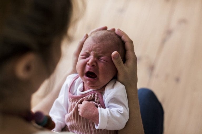 A person with long hair holds a crying newborn, possibly showing colic symptoms. The baby, dressed in a white long-sleeve shirt and pink striped overalls, is gently supported by the person's hands on their head and back. The background features a wooden floor.