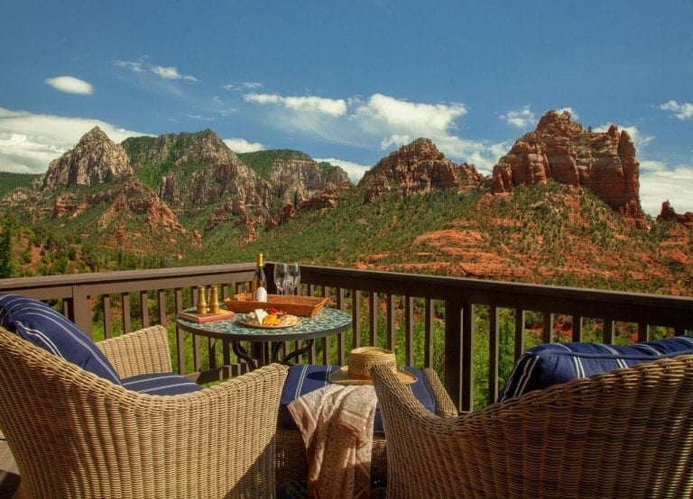 A scenic view of red rock formations and green valleys is seen from a wooden deck. The deck, perfect for a babymoon destination, has a small round table set with a bottle of wine, glasses, a plate of cheese, and two wicker chairs with cushions. A straw hat rests on one of the chairs.