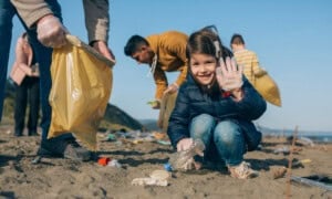 A young girl smiling and holding a plastic bottle while participating in a beach cleanup. She is wearing gloves and is surrounded by other volunteers picking up litter—one person holds a yellow bag. The sky is clear, and the beach is scattered with trash, showcasing random acts of kindness in action.