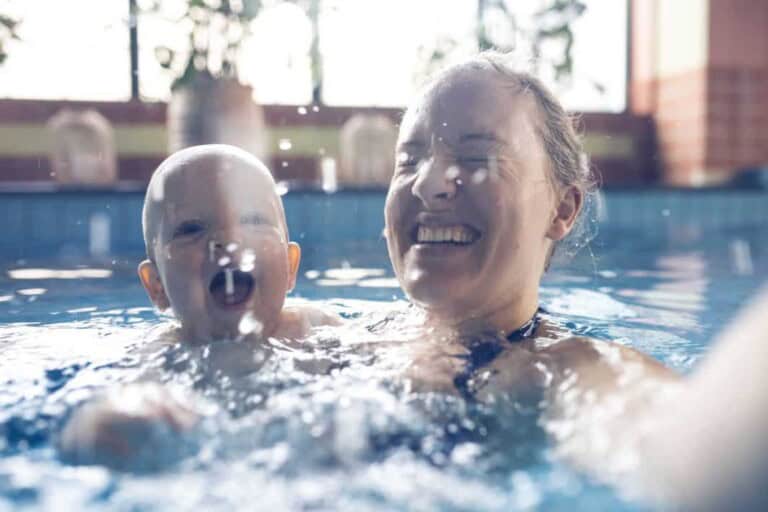 A person and a child are in a swimming pool, both smiling and enjoying the water. The indoor pool, perfect for baby swimming, has blurred windows and pots in the background. The image captures a moment of joy and playfulness.