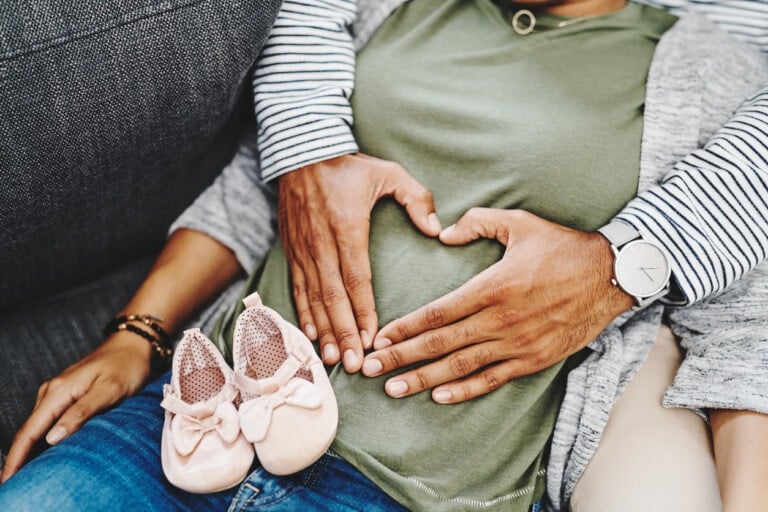Two people sitting closely, with one person's hands gently forming a heart shape on the other's pregnant belly. They are wearing casual clothes, and a pair of pink baby shoes is placed on their lap—the moment capturing not just their love but also the truths about the first trimester.