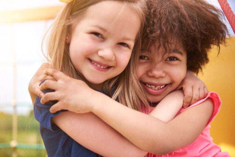 Two children smile at the camera while embracing each other, showcasing the power of words in their radiant expressions. One child has straight, blonde hair and wears a blue shirt, while the other child has curly, brown hair and wears a pink shirt. The background is bright and sunny with outdoor play equipment visible.