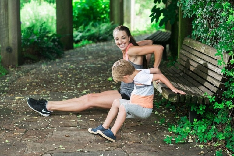 A woman and a young boy are staying active on a stone path in a shaded, wooded area. The woman is performing tricep dips on a wooden bench while the boy sits beside her on the same bench. The surroundings are filled with greenery.