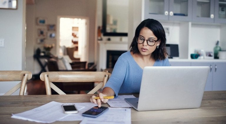 A woman with glasses is sitting at a table working on her laptop, meticulously calculating the annual family budget. She is using a calculator and has papers spread out in front of her. The background shows a cozy living room with shelves and furniture.