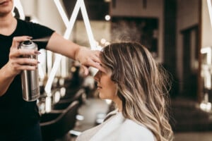 A hairdresser is applying hairspray to the hair of a woman with long, wavy hair in a salon. The woman is seated and covered with a cape, and the hairdresser is using their other hand to shield the woman's eyes from the spray. This thoughtful service makes it an ideal haircut for moms seeking comfort and style.