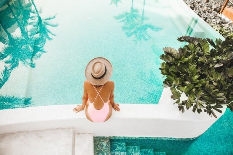 A person wearing a beige hat and a pink swimsuit that covers stretch marks sits on the edge of a pool. The pool reflects the image of palm trees. There is a plant with large green leaves on the right side of the image.