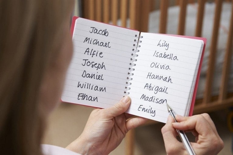 A person is holding a notebook open to a page with handwritten names, perhaps choosing a baby name. On the left page, the names listed are Jacob, Michael, Alfie, Joseph, Daniel, William, and Ethan. On the right page, the names listed are Lily, Isabella, Olivia, Hannah, Abigail, Madison, and Emily.