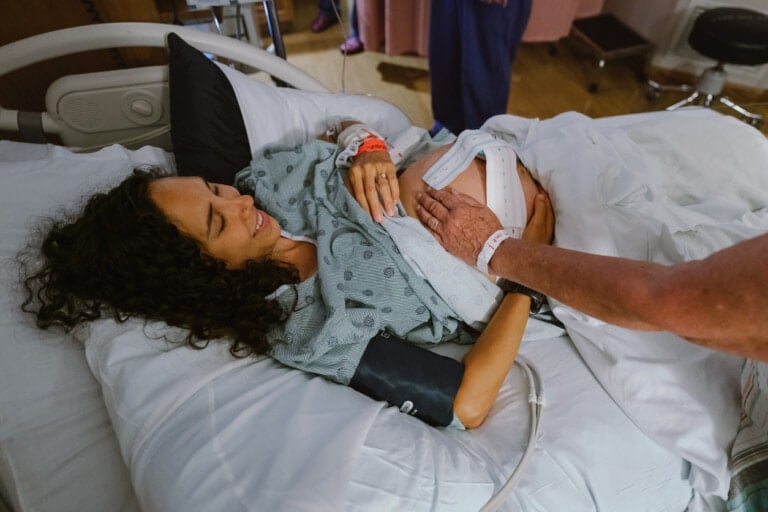 A woman is lying in a hospital bed, smiling and wearing a hospital gown. A person is placing their hand on her belly, checking on her. Medical equipment is visible in the background. The woman is also wearing an identification wristband—just in case no one told you, labor can be quite the journey!