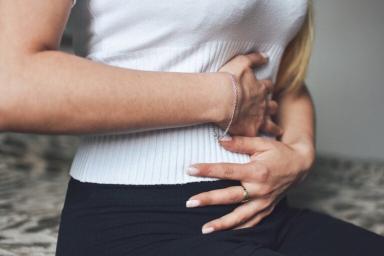 A person wearing a white shirt and black pants is sitting while holding their stomach with both hands, indicating possible discomfort or pain, which could be signs of ectopic pregnancy symptoms.
