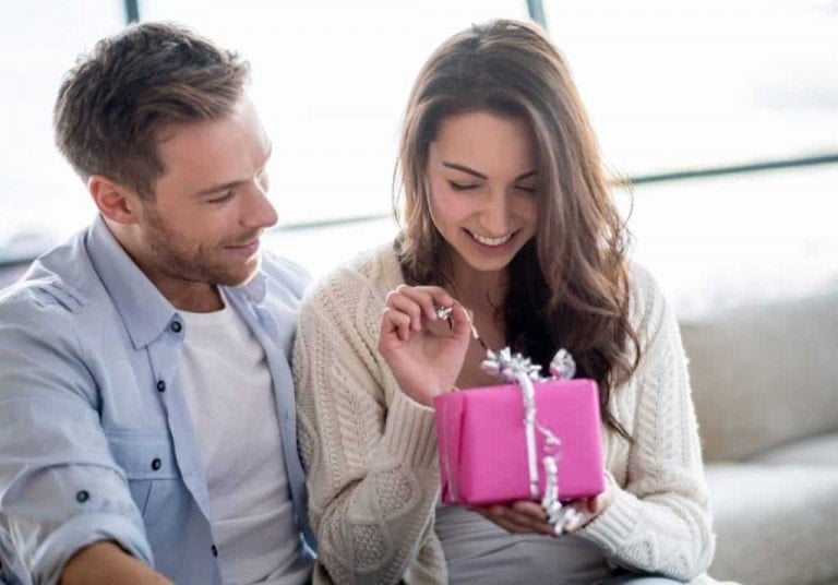 A man and a woman are sitting close together on a couch. The woman is holding a wrapped pink gift and is smiling as she unties the silver ribbon. The man, having given her this thoughtful push present, is looking at her and also smiling. They appear to be enjoying a pleasant moment.