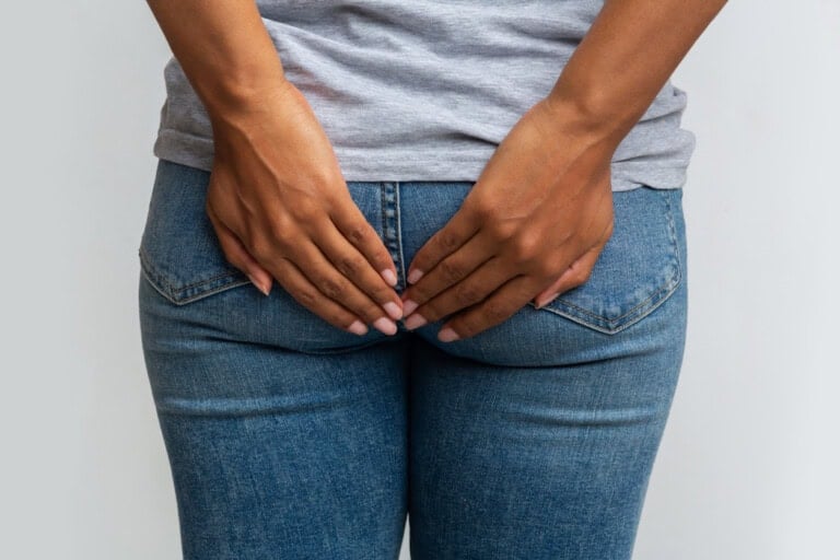 A person in a gray t-shirt and blue jeans is facing away from the camera, with hands placed at the center of the lower back, covering the buttocks—possibly practicing hemorrhoids self-care. The image shows the person from the waist down.
